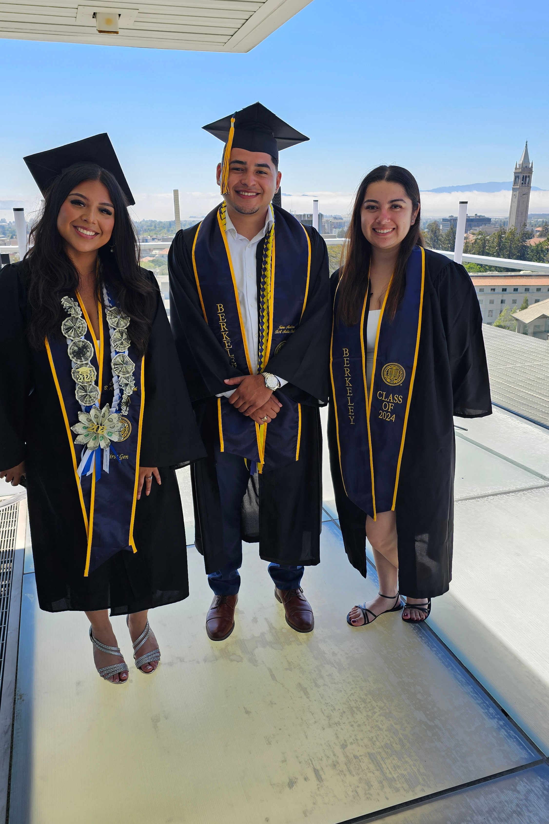 Three UC Berkeley graduates pose for a photo in cap and gown outside of the University Club with the Campanile in the background.
